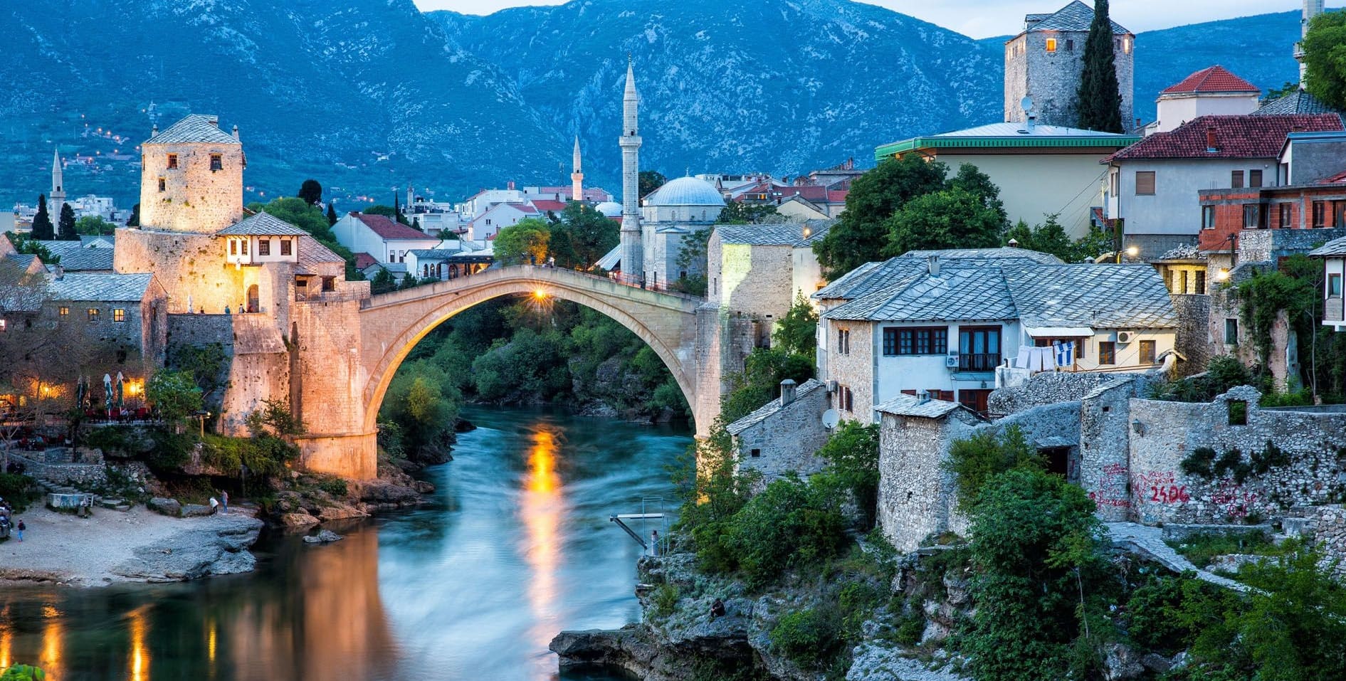 The Old Bridge of Mostar lit at dusk above the Neretva river, with a minaret and mountains behind