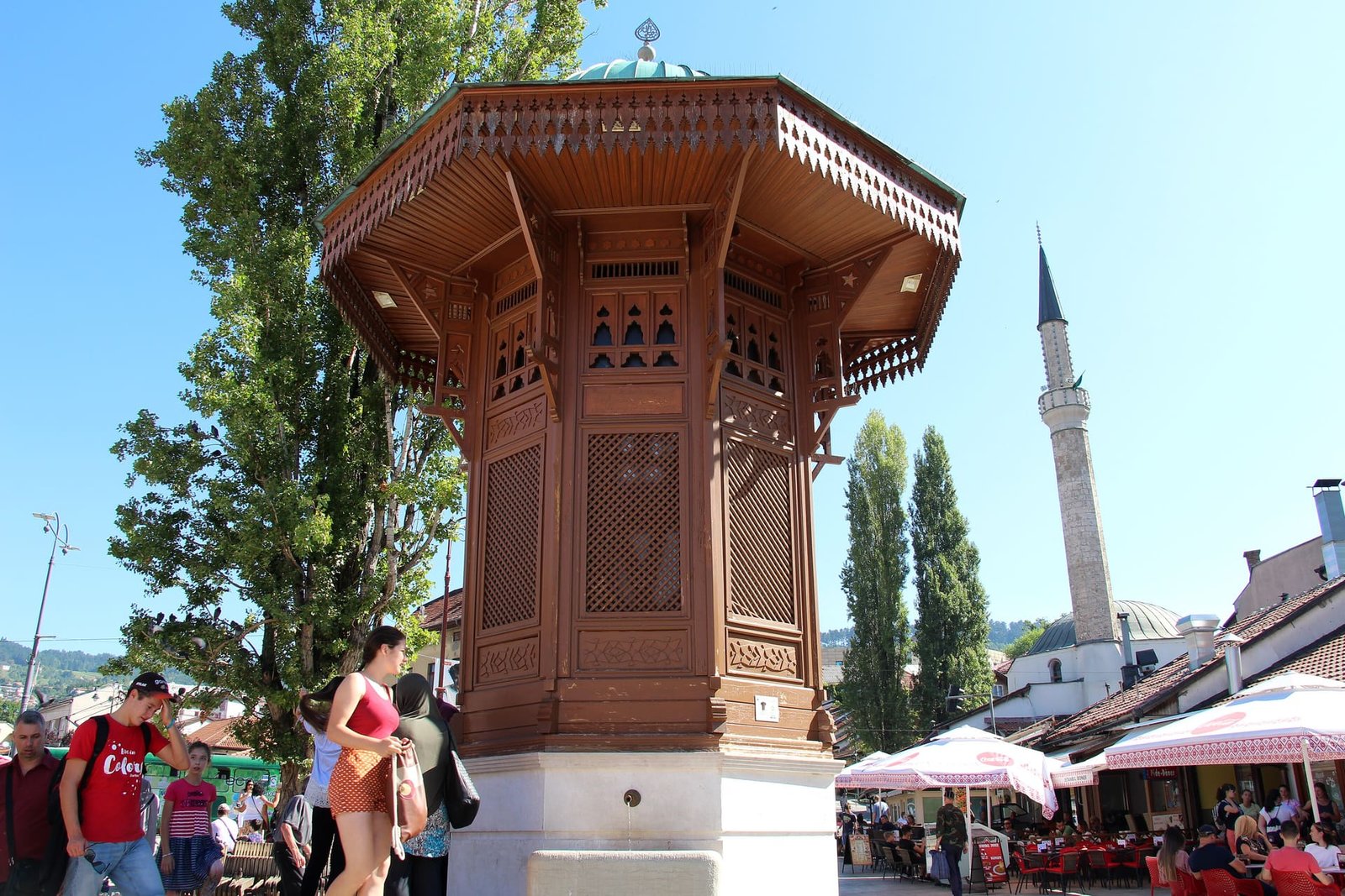 The wooden Sebilj fountain in Sarajevo's Baščaršija square with a minaret and cafe umbrellas behind it